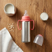 Stainless steel baby bottle with pink handles and nipple, surrounded by milk jug, glass of milk, oat flakes on wooden table